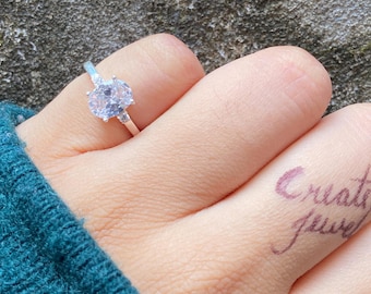 May include: A silver ring with a large, oval-shaped, clear gemstone as the centerpiece. Two smaller clear stones flank the main stone. The ring is on a finger with a tattoo that says "Create Jewel".