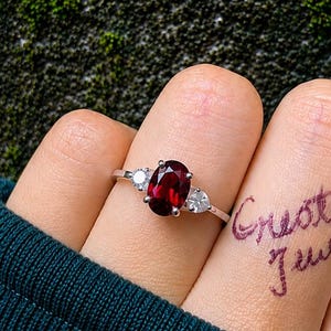 Può includere: Un anello color argento con una pietra preziosa rosso scuro dal taglio ovale al centro, affiancata da due pietre più piccole e trasparenti. L'anello è indossato su un dito con il testo scritto a mano "Create it Jewel" visibile sulla pelle.