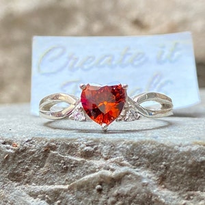 Peut inclure: Une bague en argent avec une pierre rouge en forme de cœur et de petites pierres claires sur la bande. La bague est posée sur une surface grise texturée avec une carte blanche sur laquelle est inscrit "Create it" en lettres noires.