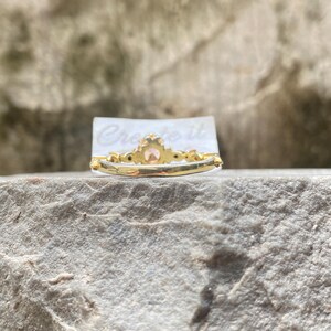 May include: A gold ring with a central peach-colored gemstone and smaller accent stones. The ring is set on a gray stone surface, with the text "Create it" visible on a white card behind the ring.