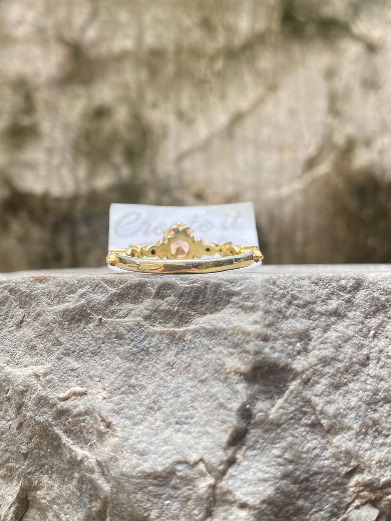 May include: A gold ring with a central peach-colored gemstone and smaller accent stones. The ring is set on a gray stone surface, with the text "Create it" visible on a white card behind the ring.