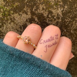 May include: A gold ring with a peach-colored gemstone is worn on a finger. The ring has a delicate design with small accent stones. The words "Create it Jewels" are written on the finger. The person is wearing a teal sweater.
