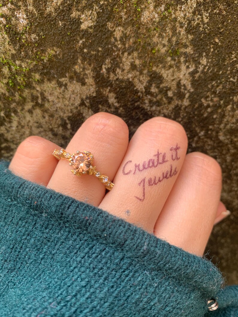 May include: A gold ring with a peach-colored gemstone is worn on a finger. The ring has a delicate design with small accent stones. The words "Create it Jewels" are written on the finger. The person is wearing a teal sweater.