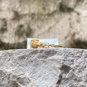 May include: A gold ring with a large, square-cut, orange gemstone and a delicate band adorned with small, clear gemstones. The ring is set on a gray, textured rock surface, with a blurred background.