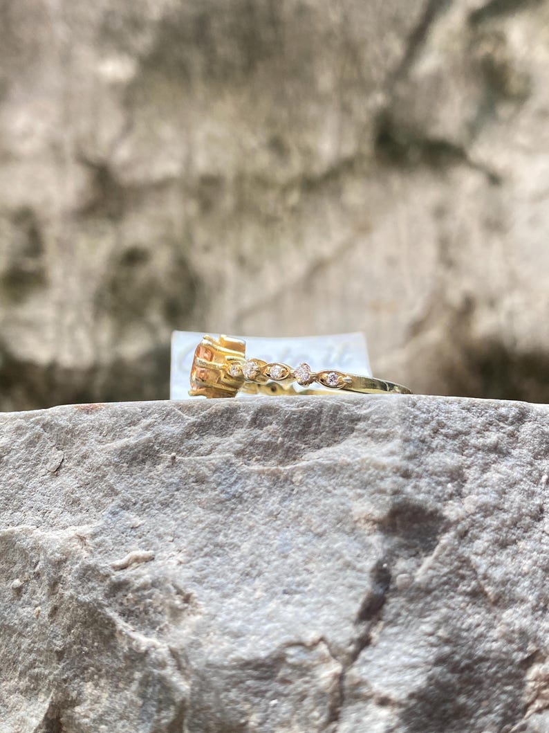 May include: A gold ring with a large, square-cut, orange gemstone and a delicate band adorned with small, clear gemstones. The ring is set on a gray, textured rock surface, with a blurred background.