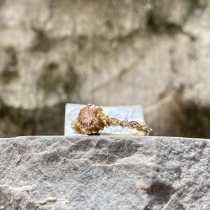 May include: A gold ring with a large, peach-colored gemstone. The band features delicate floral and diamond accents. The ring is set on a natural stone surface, showcasing its intricate design and craftsmanship. A beautiful piece of jewelry.
