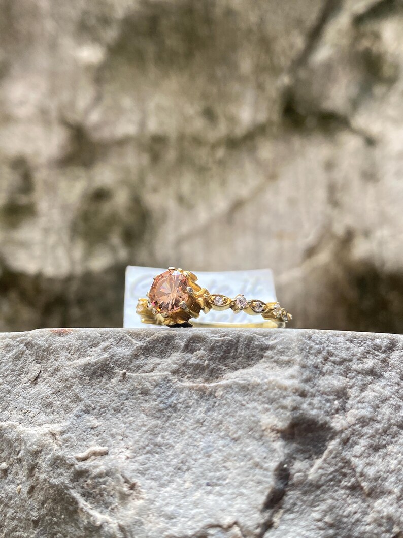 May include: A gold ring with a large, peach-colored gemstone. The band features delicate floral and diamond accents. The ring is set on a natural stone surface, showcasing its intricate design and craftsmanship. A beautiful piece of jewelry.