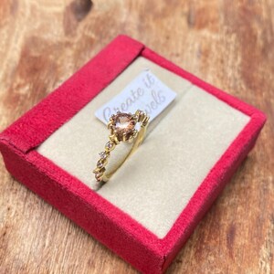 May include: A gold ring with a large, round, peach-colored gemstone at the center, surrounded by smaller clear stones. The ring is displayed in a red velvet box with the text "Create it Jewels" on a small tag.
