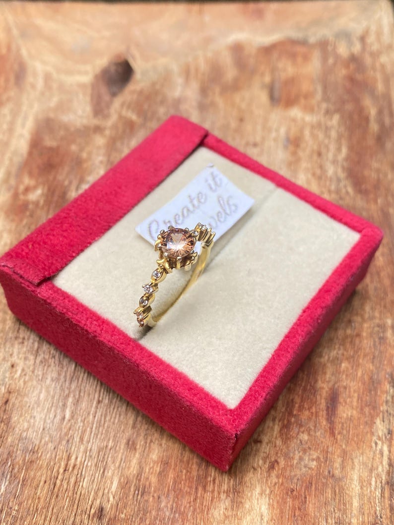 May include: A gold ring with a large, round, peach-colored gemstone at the center, surrounded by smaller clear stones. The ring is displayed in a red velvet box with the text "Create it Jewels" on a small tag.