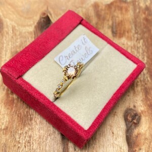May include: A gold ring with a peach-colored gemstone center and small clear stones. The ring is displayed in a red velvet box with the text "Create it Jewels" on a small tag.