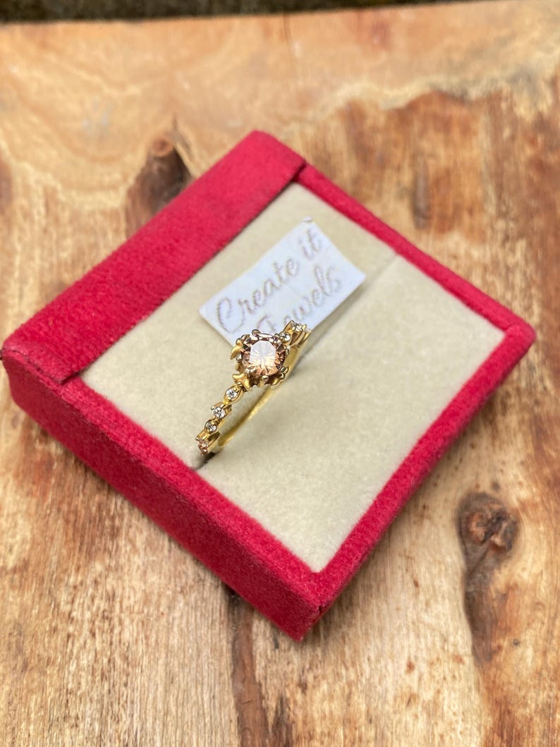 May include: A gold ring with a peach-colored gemstone center and small clear stones. The ring is displayed in a red velvet box with the text "Create it Jewels" on a small tag.