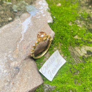 May include: A gold ring with a large, peach-colored gemstone and smaller clear stones. The ring is on a rock with a tag that reads "Create it Jewels". The background is green moss.