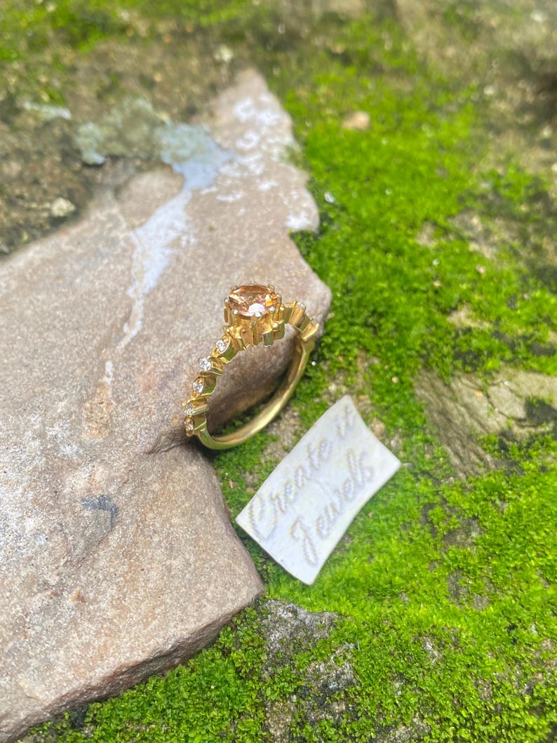May include: A gold ring with a large, peach-colored gemstone and smaller clear stones. The ring is on a rock with a tag that reads "Create it Jewels". The background is green moss.