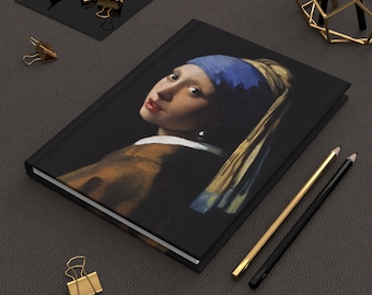 May include: A black journal featuring the painting "Girl with a Pearl Earring" by Johannes Vermeer. The cover displays the portrait of a young woman with a blue headscarf and pearl earring. Gold binder clips and pencils are arranged around the journal.