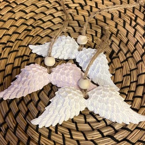 Könnte beinhalten: Drei weiße und rosafarbene Engelsflügel-Anhänger mit einer Schnur und Holzperlen, die daran hängen. Die Anhänger befinden sich auf einem geflochtenen Weidenhintergrund.