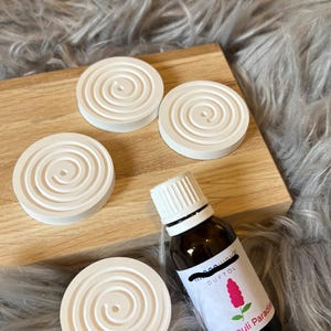 Könnte beinhalten: Vier weiße Keramik-Diffusor-Pads mit Spiraldesign und eine kleine Flasche ätherisches Öl mit der Aufschrift "Patchouli Paradise" und einer pinkfarbenen Blumengrafik.