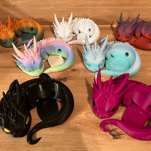 Könnte beinhalten: Sieben kleine Drachenfiguren in verschiedenen Farben, darunter Schwarz, Weiß, Rosa, Blau und Lila. Jeder Drache ist in einer schlafenden Position zusammengerollt, mit einem kleinen Loch im Rücken für eine Kerze oder ein Teelicht.