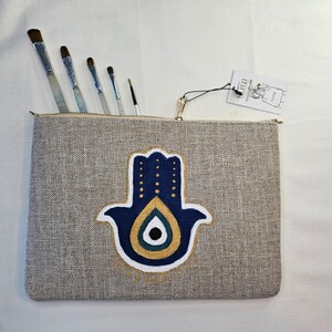 Può includere: Una pochette con zip in tela grigia con un design a mano di Hamsa blu e oro. La mano di Hamsa ha un occhio blu al centro. La pochette è decorata con una zip dorata e un piccolo cartellino che dice "Glitza".