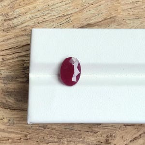 May include: A faceted, oval-shaped ruby gemstone. The gem is a deep red colour and sits on a white surface. The background is a light brown wood grain.