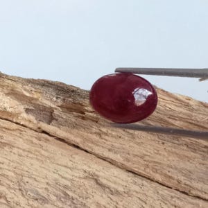 May include: A polished, oval-shaped, deep red gemstone is held by tweezers against a light brown, textured wooden surface. The gemstone reflects light, creating a bright spot on its surface. The background is a soft, light blue.