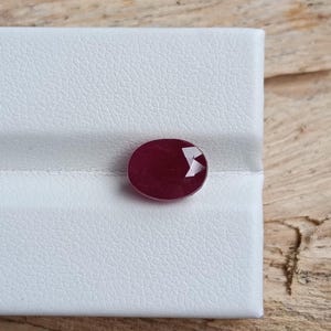 May include: A faceted, oval-shaped ruby gemstone. The deep red gem is set against a white, textured surface. The background is a light-colored wood grain.