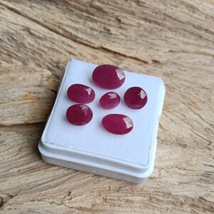 Op de afbeelding: Een verzameling van zeven gefacetteerde, ovaalvormige robijnen in verschillende maten, gepresenteerd in een witte plastic bak. De edelstenen vertonen een diepe, rijke rode kleur en zijn geplaatst tegen een houten achtergrond.