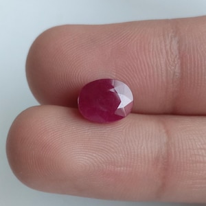 Op de afbeelding: Een gefacetteerde, ovaalvormige robijn edelsteen. De robijn heeft een dieprode kleur en een gepolijst, reflecterend oppervlak. De edelsteen wordt tussen twee vingers gehouden.