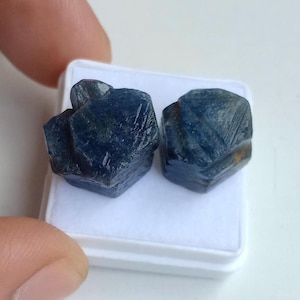 May include: Two raw, uncut sapphire gemstones. The sapphires are a deep blue color with a rough, textured surface. They are displayed on a white surface, showcasing their natural, unpolished state. The gemstones are a natural product.