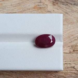 May include: A faceted, oval-shaped ruby gemstone. The gem is a deep, rich red colour with a polished surface and a cut facet on one side. The ruby is set against a white background.