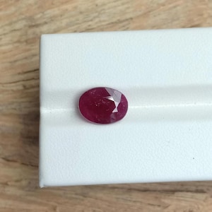 Peut inclure: Une pierre précieuse de forme ovale, un rubis facetté. Le rubis est d'un rouge profond et riche, posé sur un fond blanc. La taille de la pierre est conçue pour maximiser la réflexion de la lumière.