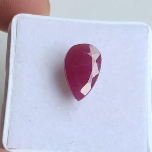 Könnte beinhalten: Ein facettierter, birnenförmiger Rubin-Edelstein. Der Edelstein hat eine tiefe, satte rote Farbe mit mehreren Facetten, die das Licht einfangen. Der Rubin ist vor einem weißen Hintergrund platziert, der seine Farbe und Form hervorhebt.