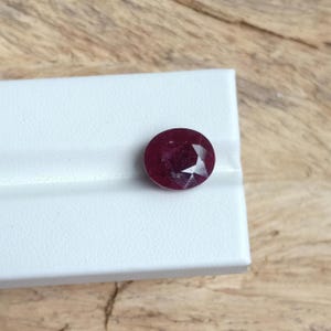 Puede incluir: Una gema de rubí facetada de forma ovalada. La gema de color rojo intenso se encuentra sobre una superficie blanca, con un fondo de madera. El corte del rubí revela múltiples facetas, realzando su brillo y color.