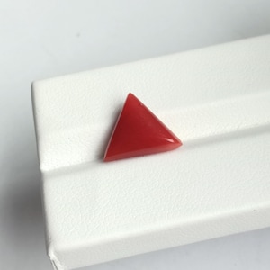 May include: A vibrant red, triangular gemstone, possibly coral, rests on a white textured surface. The gemstone's shape is a perfect equilateral triangle, showcasing a polished, smooth surface. The color is a deep, saturated red.