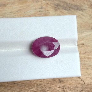 May include: A faceted, oval-shaped ruby gemstone. The gem is a deep, rich red color and sits on a white surface. The gemstone's cut reveals multiple facets, reflecting light.