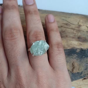 Pode incluir: Um anel de prata com uma grande pedra de aguamarina em bruto. A pedra azul-esverdeada clara tem uma forma irregular e é presa por quatro garras. O anel é mostrado em uma mão.
