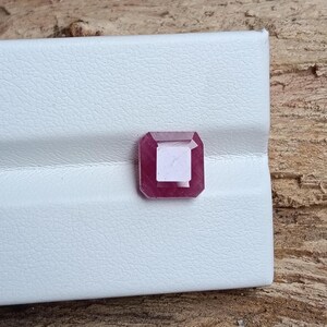 May include: A square-cut, deep red gemstone with a lighter center, set against a white textured surface. The gemstone's facets catch the light, showcasing its clarity and color. The gem is likely a ruby or similar precious stone.