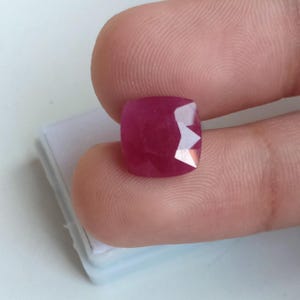 Op de afbeelding: Een gefacetteerde, kussenvormige robijn edelsteen. De robijn heeft een diepe, verzadigde rode kleur met een gepolijst oppervlak en scherpe, geometrische facetten. De edelsteen wordt tussen vingers tegen een witte achtergrond gehouden.