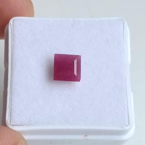 May include: A small, square-cut ruby gemstone, displayed on a white background. The ruby is a deep, saturated red color with a polished surface. The gemstone is set in a white display case.