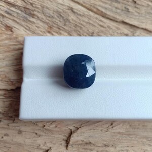 Op de afbeelding: Een donkerblauwe, ovaalvormige edelsteen met meerdere facetten. De edelsteen ligt op een wit oppervlak, met een houten achtergrond. De slijping van de steen creëert een sprankelend effect. Deze edelsteen kan in sieraden worden gebruikt.