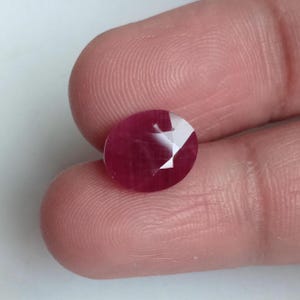Może przedstawiać: Fasetowany, owalny kamień szlachetny rubinu. Rubin ma głęboki czerwony kolor z jasnymi, odbijającymi światło fasetami. Kamień szlachetny jest trzymany między dwoma palcami, ukazując jego rozmiar i blask. Tło jest w miękkim, neutralnym odcieniu.