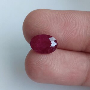 Puede incluir: Una gema de rubí facetada de forma ovalada. La gema es de un color rojo intenso y rico con una superficie pulida. Las facetas captan la luz, creando un efecto brillante. El rubí se sostiene entre dos dedos.