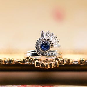 Op de afbeelding: Close-up van twee ringen op een goudkleurig decoratief dienblad. De bovenste ring heeft een blauwe middensteen en een waaiervormig ontwerp met heldere stenen. De tweede ring is een eenvoudige band. De lade heeft een gedetailleerde rand.