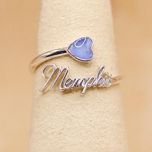 Puede incluir: Un anillo abierto de tono plateado que presenta el nombre Memphis en una fuente cursiva en un extremo y un pequeño acento en forma de corazón de color violeta claro en el otro. El anillo tiene un acabado metálico pulido y un diseño abierto.