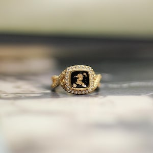 Op de afbeelding: Een gouden ring met een vierkante zwarte steen gezet in een ruitvormig frame. De ring heeft een gedraaide band.