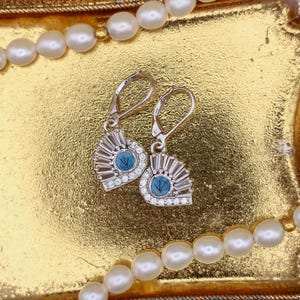 Op de afbeelding: Zilverkleurige oorbellen met een waaiervormig ontwerp, met een centrale blauwe steen omringd door kleine, heldere stenen. De oorbellen worden gepresenteerd op een goudkleurig dienblad, met een parelketting op de achtergrond. De oorbellen hebben een klapbeugel sluiting.