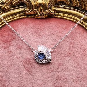 Op de afbeelding: Een zilveren ketting met een hanger met een centrale blauwe edelsteen omringd door heldere, baguette-geslepen en ronde stenen. De ketting wordt getoond tegen een roze fluwelen achtergrond en een goudkleurig, sierlijk frame.