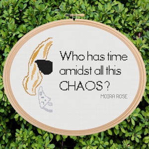 May include: White cross-stitch embroidery hoop with a quote from the TV show Schitt's Creek: "Who has time amidst all this CHAOS? Moira Rose". The embroidery features a cartoon of Moira Rose with her signature blonde hair and sunglasses.