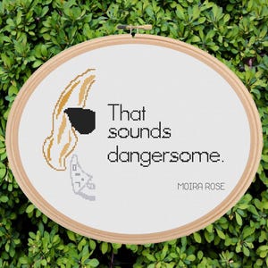 May include: Oval cross-stitch embroidery hoop with the text "That sounds dangersome." and "Moira Rose." The design includes a stylized blonde hairstyle with a black mask and a small gray object. The background is green foliage.