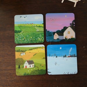 May include: A set of four square coasters, each with a different rural landscape scene. The coasters show a green field, a sunset, a harvest scene, and a snowy winter landscape. The coasters are made of a durable material.