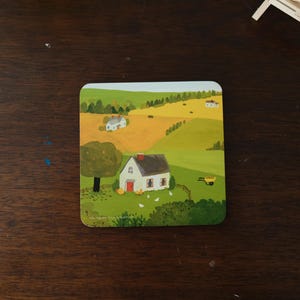 Op de afbeelding: Vierkante onderzetter met een landelijke landschapsschilderij. Het kunstwerk toont een wit huis met een rode deur en schoorsteen, omringd door groene velden en bomen. Een gele kruiwagen staat in de scène. De onderzetter ligt op een donker houten oppervlak.